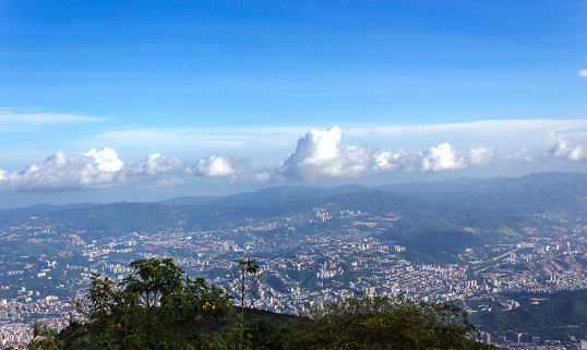 View of Caracas from the top of the Waraira Repano\nWide view of Caracas from the heights\nBlue sky with white clouds, line of mountains in the background bordering on the sky\nYou can see in the middle the beautiful city of Caracas (the valley)