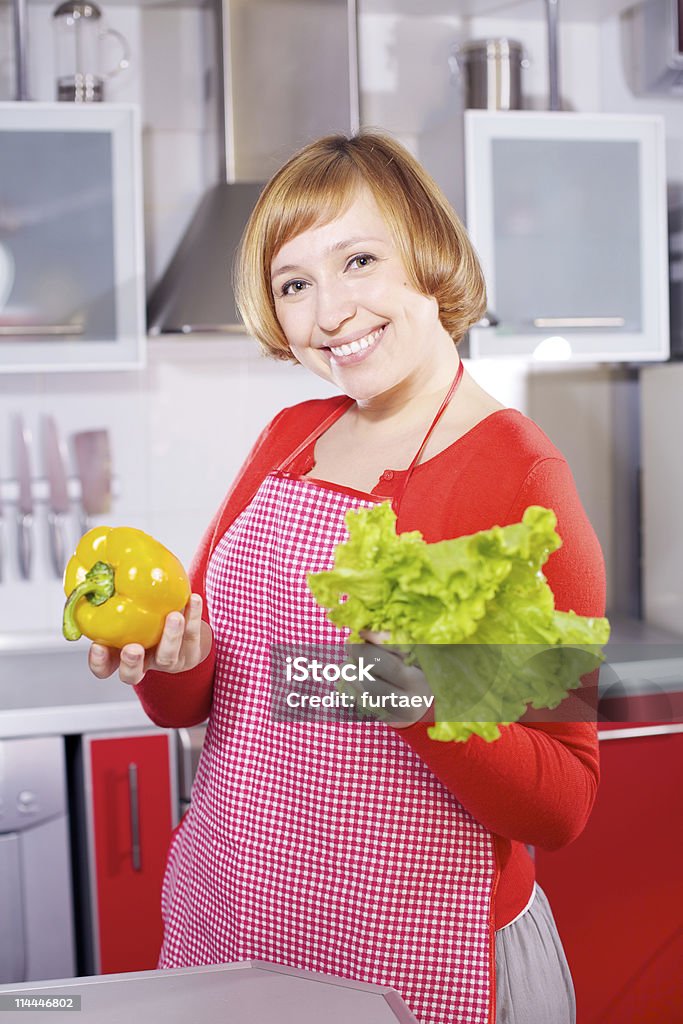 Hermosa joven ama de casa de cocina sosteniendo pimienta roja y lechuga - Foto de stock de A cuadros libre de derechos