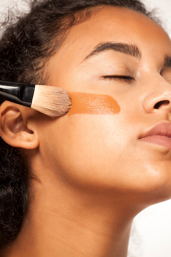 portrait of a young dark-skinned woman applying liquid makeup base with brush on her face on a white background