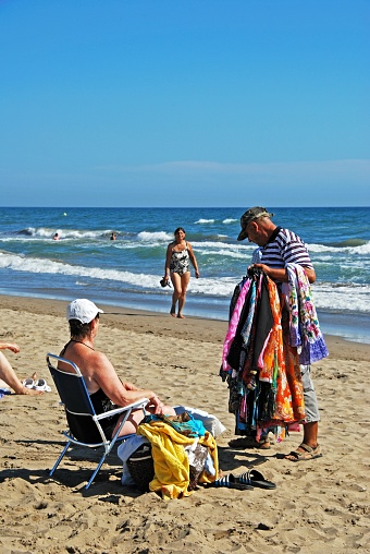 Beach trader showing his goods to a tourist on Playa de la Vibora beach, Elviria, Marbella, Malaga Province, Andalusia, Spain, Western Europe.