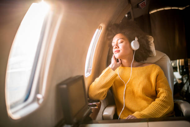 joven mujer de moda sentada en un jet privado y escuchando música a través de los auriculares. ella está mirando a través de la ventana - status symbol audio fotografías e imágenes de stock