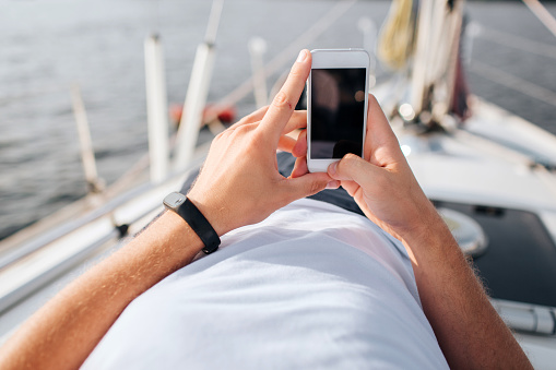 Picture of phone in man's hands. He holds in confident. Screen is dark. Phone is white. Young man lies on yacht board