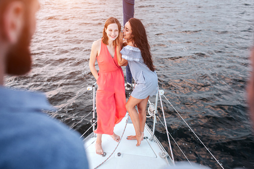 Beautiful young women stand together and pose. They wispering and laughing. Men stands adn look at them. Women are on bow of yacht