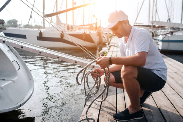 foto del hombre en gorra blanca y camisa sentada en posición de escuadrón en el muelle. tiene muchas cuerdas. el tipo es tranquilo y pacífico. está concentrado. - moored boats fotografías e imágenes de stock