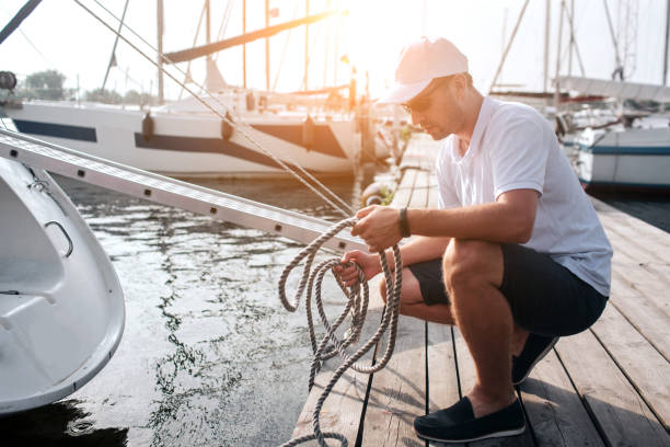 picture of man in white cap and shirt sitting in squad position on pier. he holds lots of ropes. guy is calm and peaceful. he is concentrated. - moored boats imagens e fotografias de stock