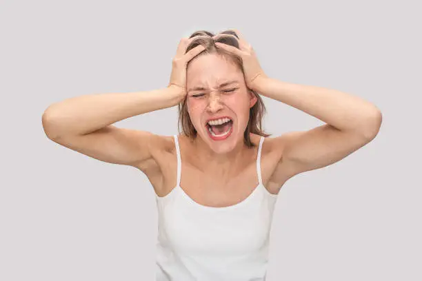 Photo of Poor young woman stands and suffering from terrible headache. She screams. Model keeps eyes closed. She wears white t-shirt. Isolated on grey background.
