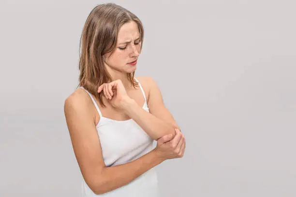 Photo of Young woman with freckles holds hand on elbow. There is pain. She is serious. Young woman wears white t-shirt. Isolated on grey background.