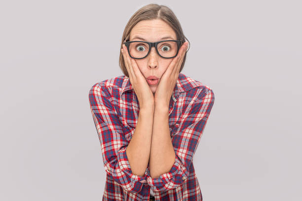istock Amazed and excited young model looks on camera though glasses. She holds hands on cheeks. Young woman is wondered. Isolated on grey background. 1144512457