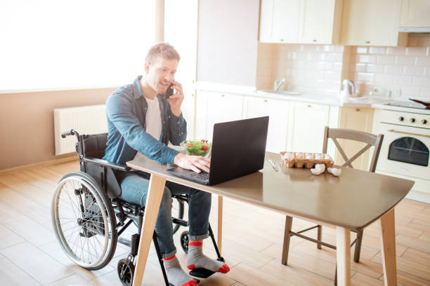 istock Young worker with disability and special needs sit at table and work. He use laptop and talk on phone. Alone in kitchen. Serious and concentrated. 1144516302