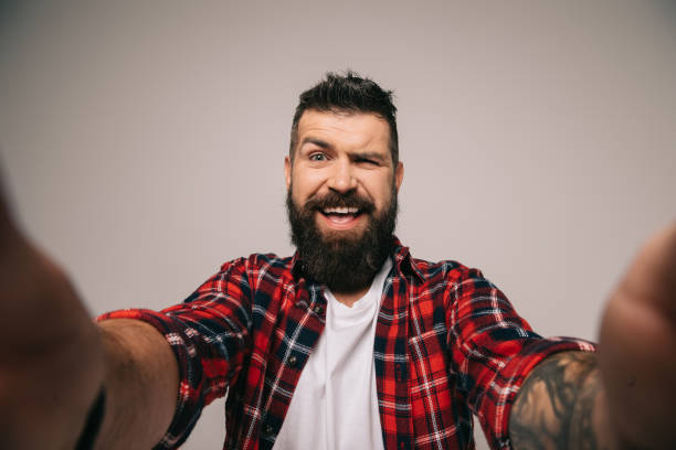 istock cheerful bearded man winking and taking selfie, isolated on grey 1144525478