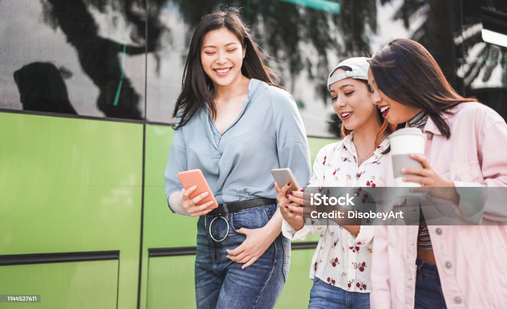 Happy asian friends using smartphones at bus station - Young students people having fun with technology trends after school - Friendship and transports app concept - Focus on center girl face Asian and Indian Ethnicities Stock Photo