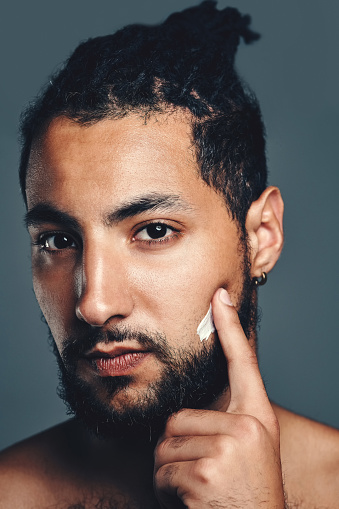 Studio portrait of a handsome young man applying moisturiser to his face against a grey background