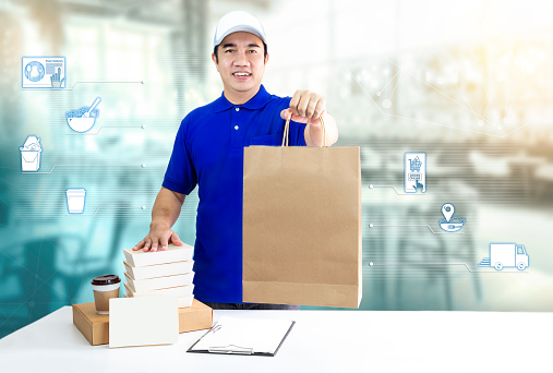 Food delivery service for order online and icon media. Delivery man in blue uniform hand holding paper bag, packaging container and disposable cup for express delivery on restaurant background.