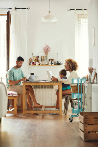Parents working while sitting with son at home Man using digital tablet while working from home. Woman using laptop with son at table. They are sitting in kitchen. family photo on wall stock pictures, royalty-free photos & images