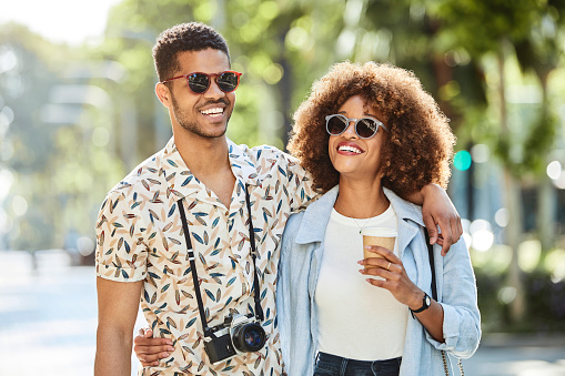Smiling young couple walking arm around on sidewalk. Male and female tourists are with camera and disposable coffee cup traveling in city. They are wearing casuals.