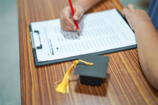 istock Asian children young kid write answer in a quiz examination test study learn with graduation gap hat on wooden table in school : bright genius education concept. 1144544025