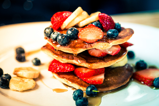 Close up color image depicting a stack of freshly cooked breakfast pancakes topped with fresh fruit - sliced banana, blueberries, strawberries - and drizzled with maple syrup. White plate in the background allows room for copy space.
