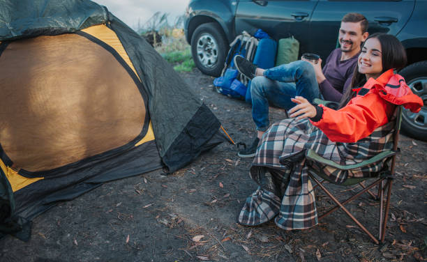 istock Beautiful and cheerful young woman points on left. They looks with guy in the same direction. People hold thermocups in hands. They sits on folding chairs at tent and car. 1144546224