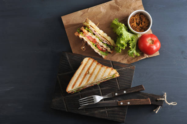 Half a club sandwich on a wooden board. View from above. Half a club sandwich on a wooden board. The other half of the sandwich is next to kraft paper. In the frame cutlery, mustard, lettuce, tomato. Dark background. Close-up. View from above. sandwich club sandwich lunch restaurant stock pictures, royalty-free photos & images