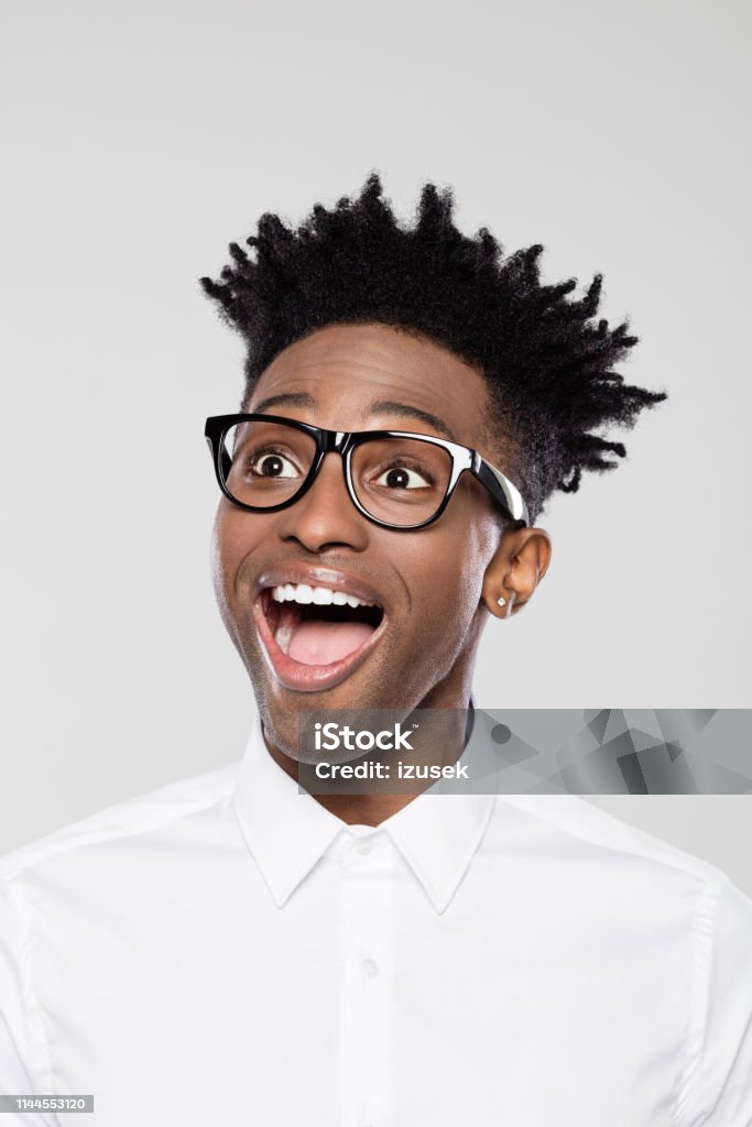 Nerdy young man looking away Close up portrait of excited afro american young businessman on gray background. Man in white shirt wearing eyeglasses with funky hairstyle looking away. Surprise Stock Photo