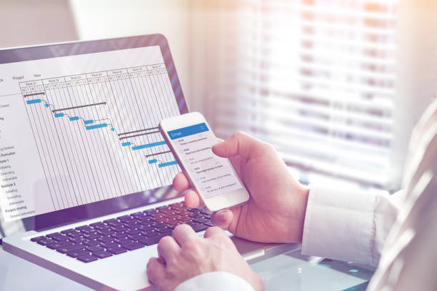 istock Project manager working on Gantt chart planning schedule with tasks and milestones on computer and sending email on smartphone, professional management and communication app, office interior 1144557364
