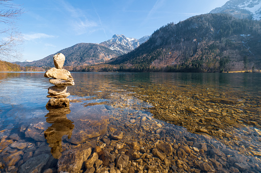 Vorderer Langbathsee near Ebensee, Oberösterreich, Austria - 30th March 2019. The forest and the mountains of the Salzkammergut reflecting on the turquoise colored water of the Vorderer Langbathsee. Some stones put together forming a manikin standing in the shallow water.