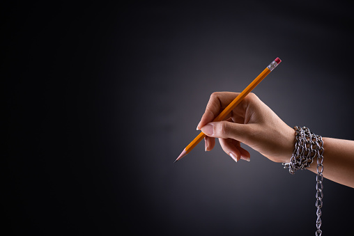 Woman hand with yellow pencil tied with chain, depicting the idea of freedom of the press or freedom of expression on dark background in low key. World press freedom and international human rights day concept.