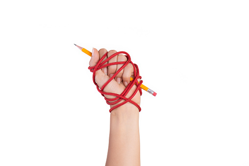 Woman hand with yellow pencil tied with red rope, depicting the idea of freedom of the press or freedom of expression on dark background in low key. World press freedom and international human rights day concept.