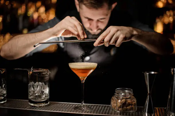 Professional bartender adding to a cocktail in the glass with a whipped cream a grated nutmeg on the bar counter in the dark blurred background.