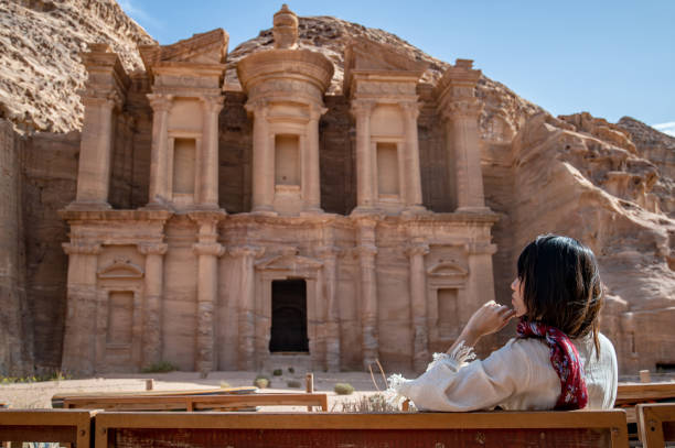 mujer asiática turista en vestido blanco sentado y mirando a ad deir o el deir, el monumento tallado en la roca en la antigua ciudad de petra, jordania. viaje patrimonio de la humanidad por la unesco en oriente medio - petra antiquities jordan middle east fotografías e imágenes de stock