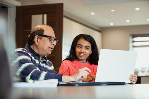 A grandfather is helping his granddaughter as she works from a laptop; they look happy to be bonding and working together.
