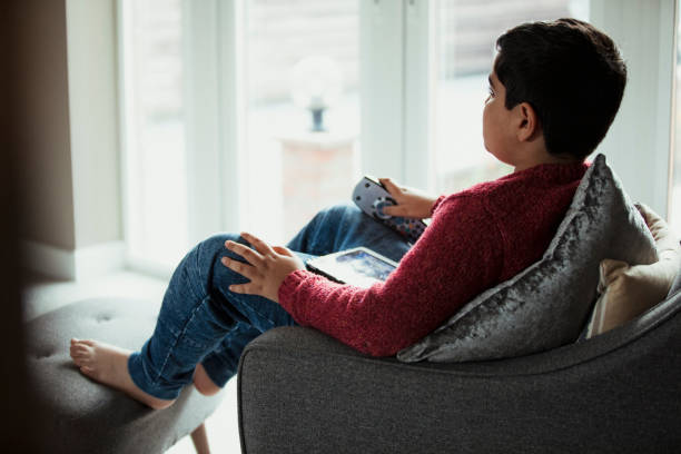 Time Alone to Relax A young boy relaxing whilst sitting on an armchair, holding a portable tablet and watching an autistic friendly program. indian boy barefoot stock pictures, royalty-free photos & images