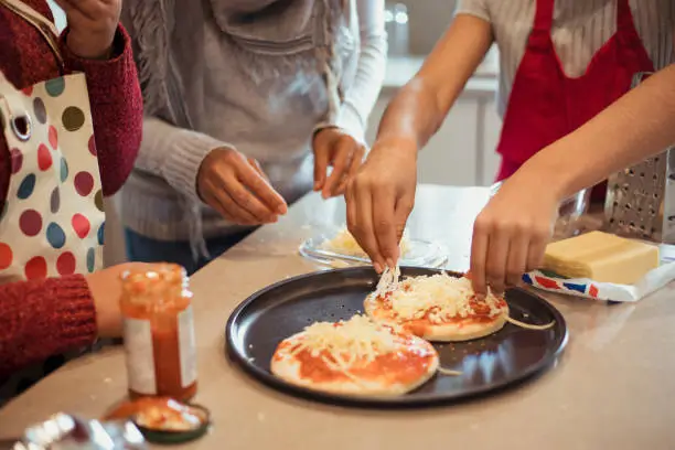 A close-up of a family including a mother, daughter and son, preparing pizzas for dinner by adding the cheese on top.