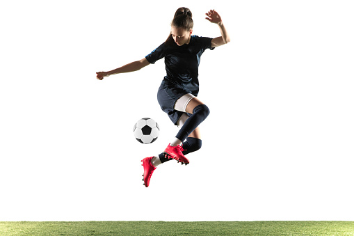 Young female soccer or football player with long hair in sportwear and boots kicking ball for the goal in jump isolated on white background. Concept of healthy lifestyle, professional sport, hobby.