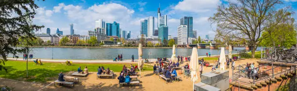 Crowds of locals and tourist enjoying the spring sunshine at an al fresco promenade cafe on the River Main waterfront overlooked by the futuristic skyscraper skyline of central Frankfurt, Hesse, Germany.