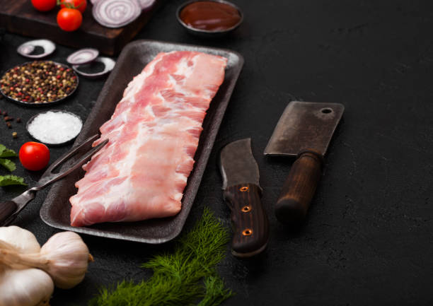 istock Plastic tray with raw pork ribs and vintage knife and fork on black stone background. Fresh tomatoes and red onion with garlic salt and pepper. 1144596385