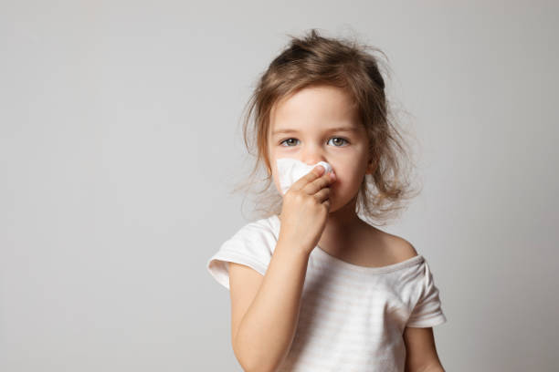 Little Girl Cleaning Her Nose Little sick girl is cleaning her nose with a white handkerchief and is looking at camera in front of a gray blank wall. blowing nose photos stock pictures, royalty-free photos & images