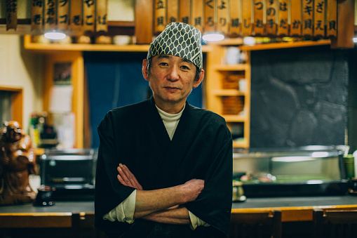 Portrait of a real Japanese Sushi Chef in Tokyo, Japan. The man is folding his arms and looking at the camera while standing in his family owned sushi restaurant.
