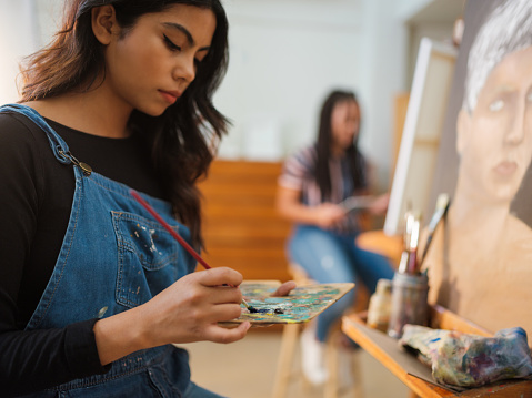 A latin female art student holding a paint palette and mixing colors with a paintbrush as she sits in front of her painting.