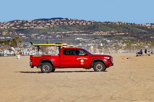 Newport Beach, CA / USA – April 6, 2019:  The City of Newport Beach’s Fire Department Lifeguard truck on sand at the Balboa Peninsula with hillside homes in the background.