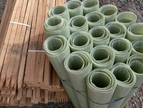 Bundles of upright green corrugated plastic tree guards stacked inside each other in groups of four or five. They are all tied together with a single thin metal strap. Beside them are stacks of long, wooden tree stakes. They are held together by a white plastic strap. The tree planting equipment is stored outside on stony ground.