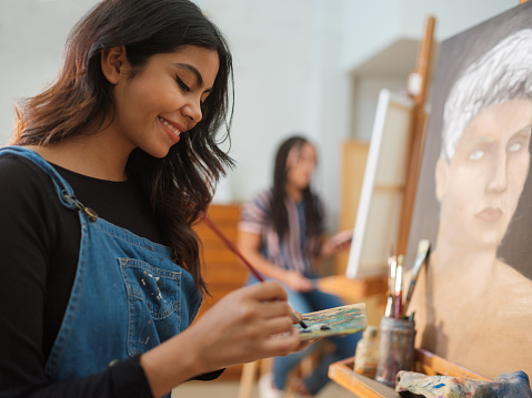 A latin female smiling as she uses a paintbrush to mix colors on a palette while standing in front of her painting.