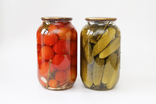 two glass jars with tomatoes and cucumbers closed with a metal lid on light background