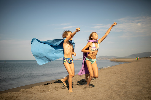 Children playing superheroes on beautiful sand beach. They wears swimwear and towels as a cape and looks very playful and happy