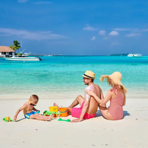 Family on beach, young couple with three year old boy. Woman applying sun screen protection lotion on man's back. Summer vacation at Maldives.