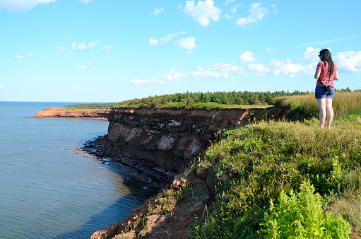 A woman stands on the edge of the famous red cliffs on Prince Edward Island.  She is looking out at the ocean. Prince Edward Island National Park. Summer. Day. Circa 2012.