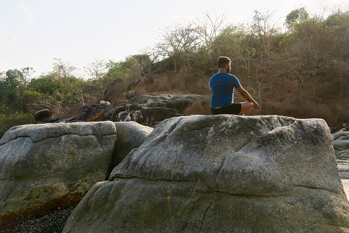 Rear view of Indian man wearing shorts and a blue t-shirt practicing Lotus Pose (Padmasana) yoga position on top of a rock at the water's edge of Palolem Beach, Goa, India.