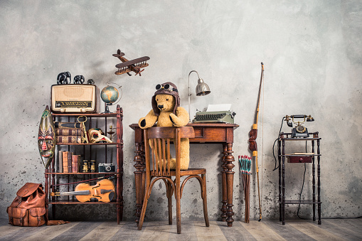 Retro Teddy Bear toy on chair, typewriter, desk lamp, vintage telephone on stand, old books, radio, globe,  binoculars, carnival mask, photo camera, fiddle on shelf, wooden plane, travel backpack, bow