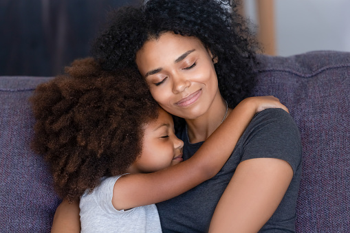 Close up biracial family portrait loving mother and little daughter sitting on couch at home hugging with closed eyes. Love, new mom for adopted child, warm relationships, caring elder sister concept