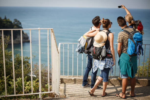 Four young people are posing for a selfie while exploring the coastline on summer vacation. They are enjoying beautiful view of sea against sky.